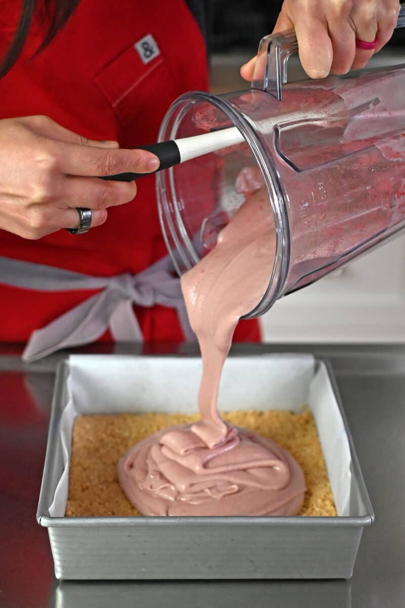 Pouring the pink vegan strawberry cheesecake filling onto the crust in a lined baking pan.