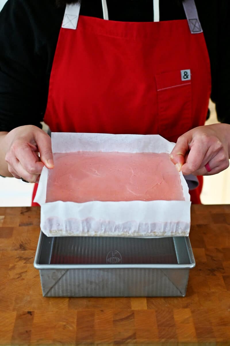 Removing the chilled strawberry cheesecake bars from the pan by pulling up on the parchment paper.