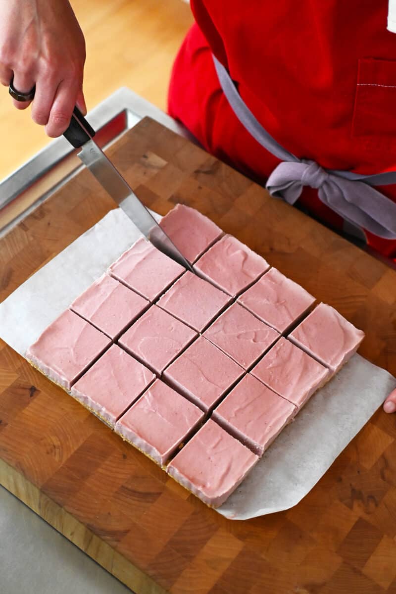 An overhead shot of someone cutting a batch of vegan and paleo strawberry cheesecake bars into 16 square pieces.