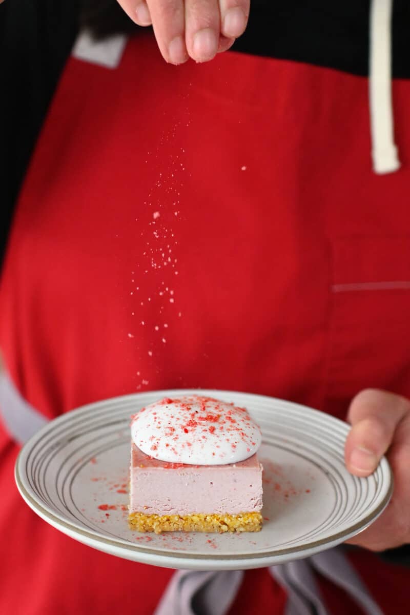 A person in a red apron is holding a plate with a strawberry cheesecake bar topped with whipped coconut cream and strawberry powder.