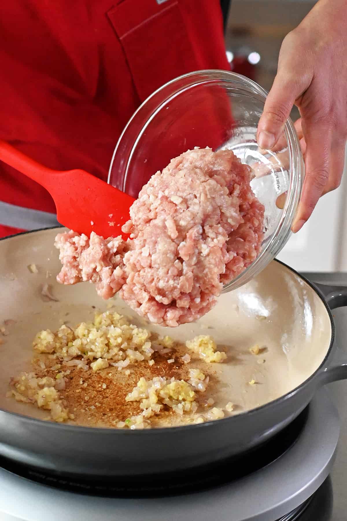 Adding raw ground chicken to a skillet with minced shallots, garlic, and ginger.