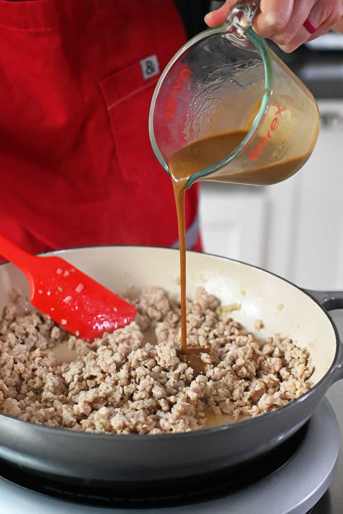 Pouring a brown sauce from a measuring cup into a skillet with cooked ground chicken.