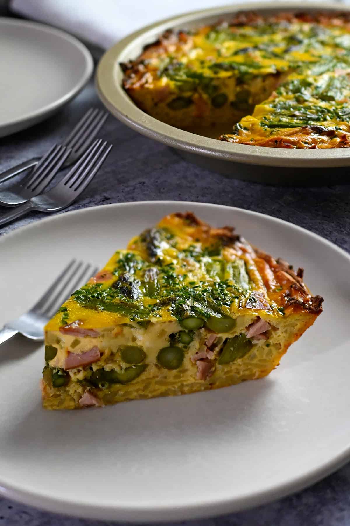 A slice of paleo and dairy free asparagus quiche on a plate in front of pie pan with quiche inside.
