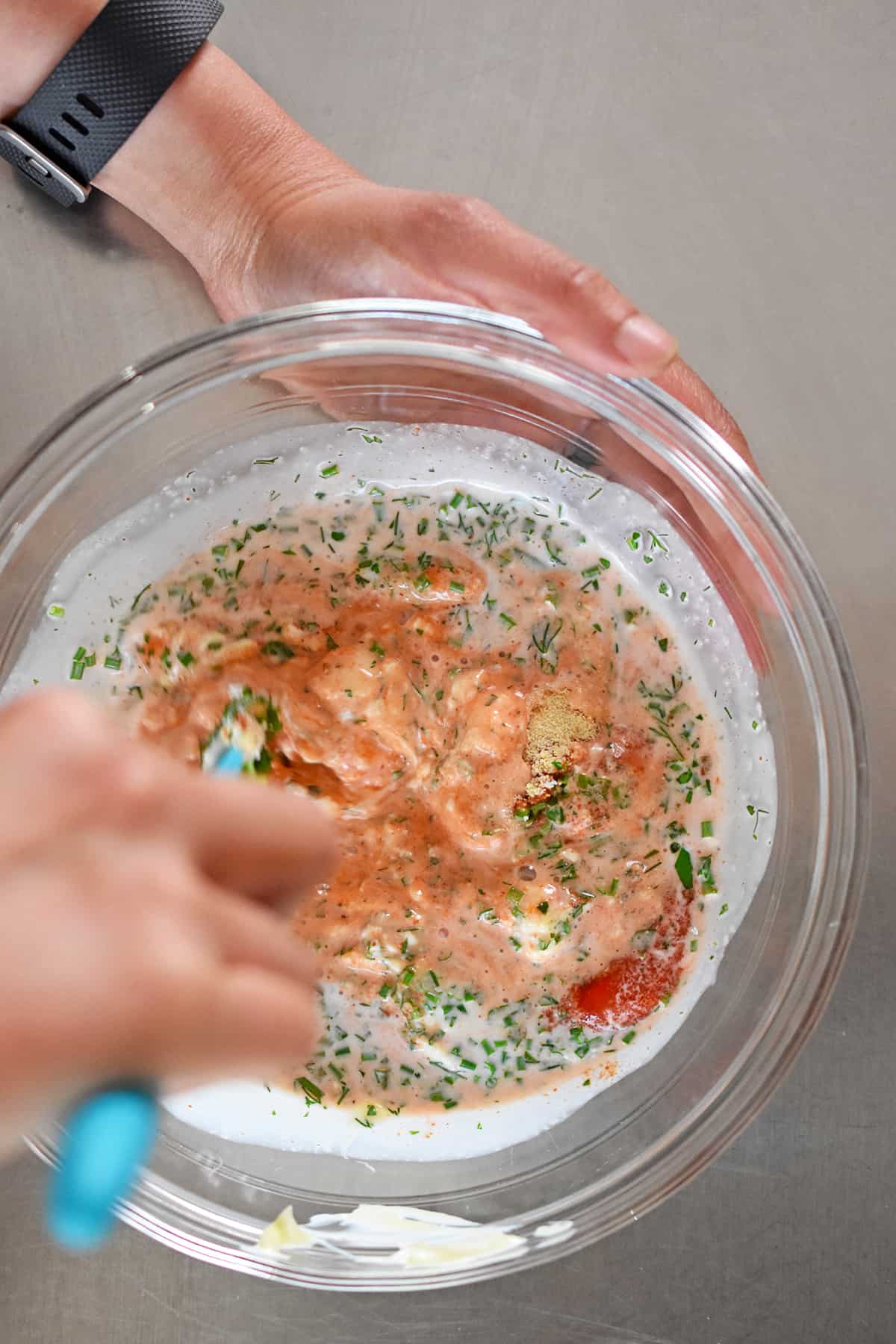 An overhead shot of someone using a blue silicone spatula to mix a bowl of paleo sriracha ranch dressing.