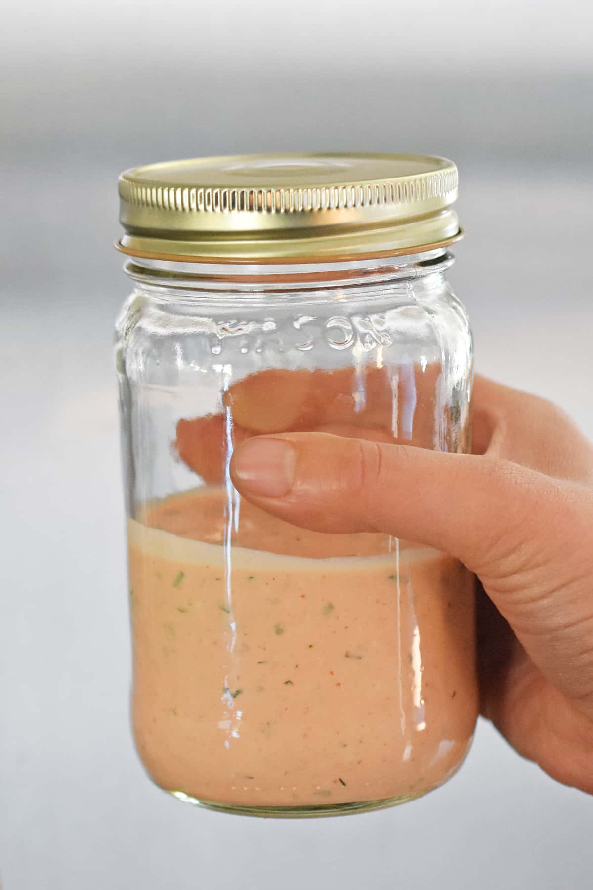 A hand is holding a sealed glass mason jar filled with creamy sriracha ranch dressing.