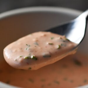 A spoon is scooping some creamy orange homemade sriracha ranch dressing in a white bowl.