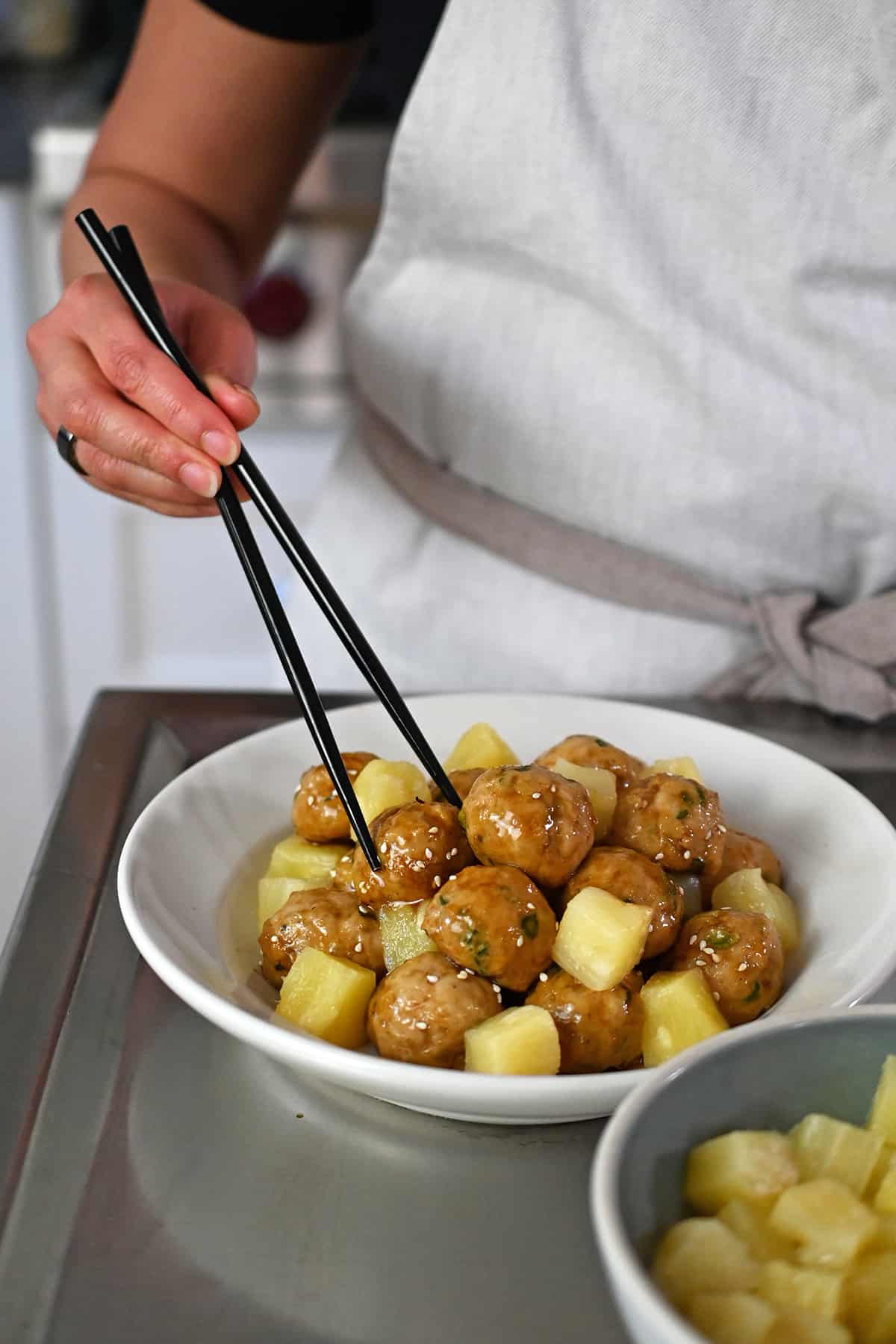 A pair of chopsticks is grabbing a teriyaki pineapple meatball from a bowl with cubed pineapple.