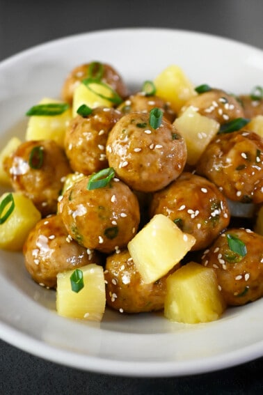 A side view of a plate of teriyaki pineapple chicken meatballs with cubed pineapple.