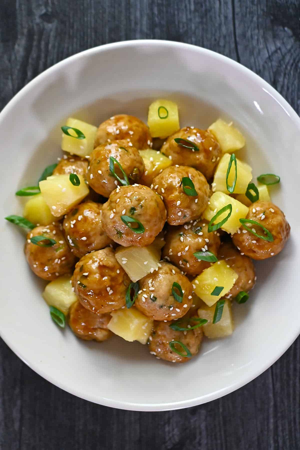 An overhead shot of a white plate filled with teriyaki pineapple meatballs and cubed pineapple, topped with toasted sesame seeds and sliced green onions.