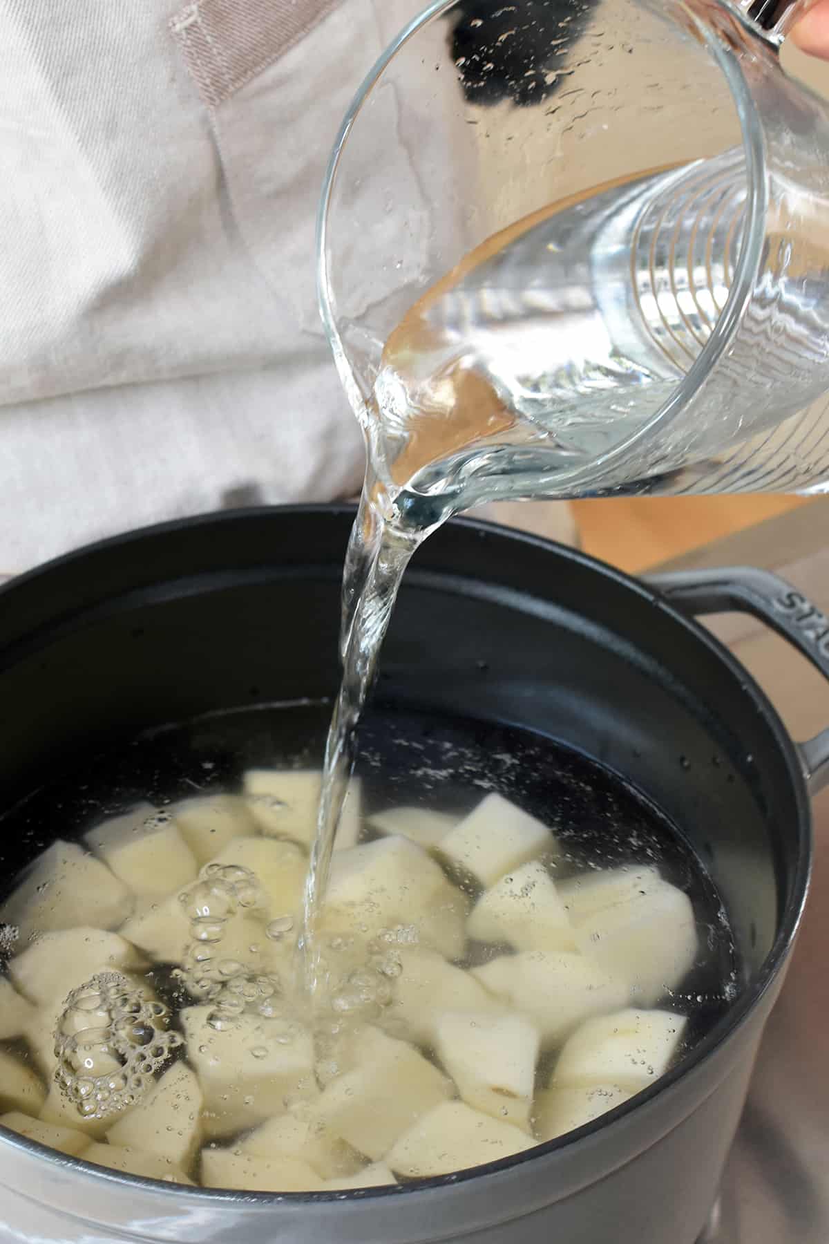 Pouring water into a large pot filled with cubed raw russet potatoes.