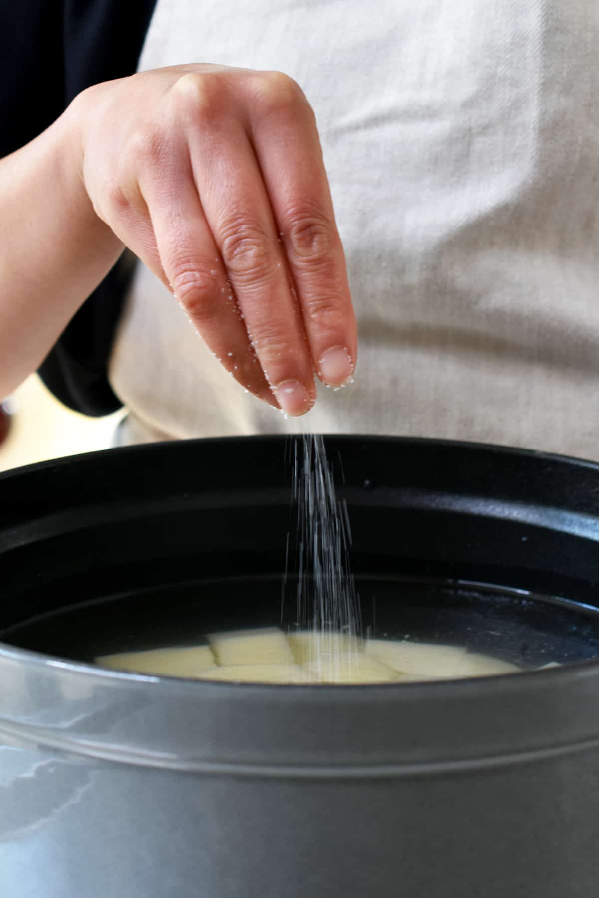 A hand is sprinkling salt into a pot filled with cubed raw potatoes and water.