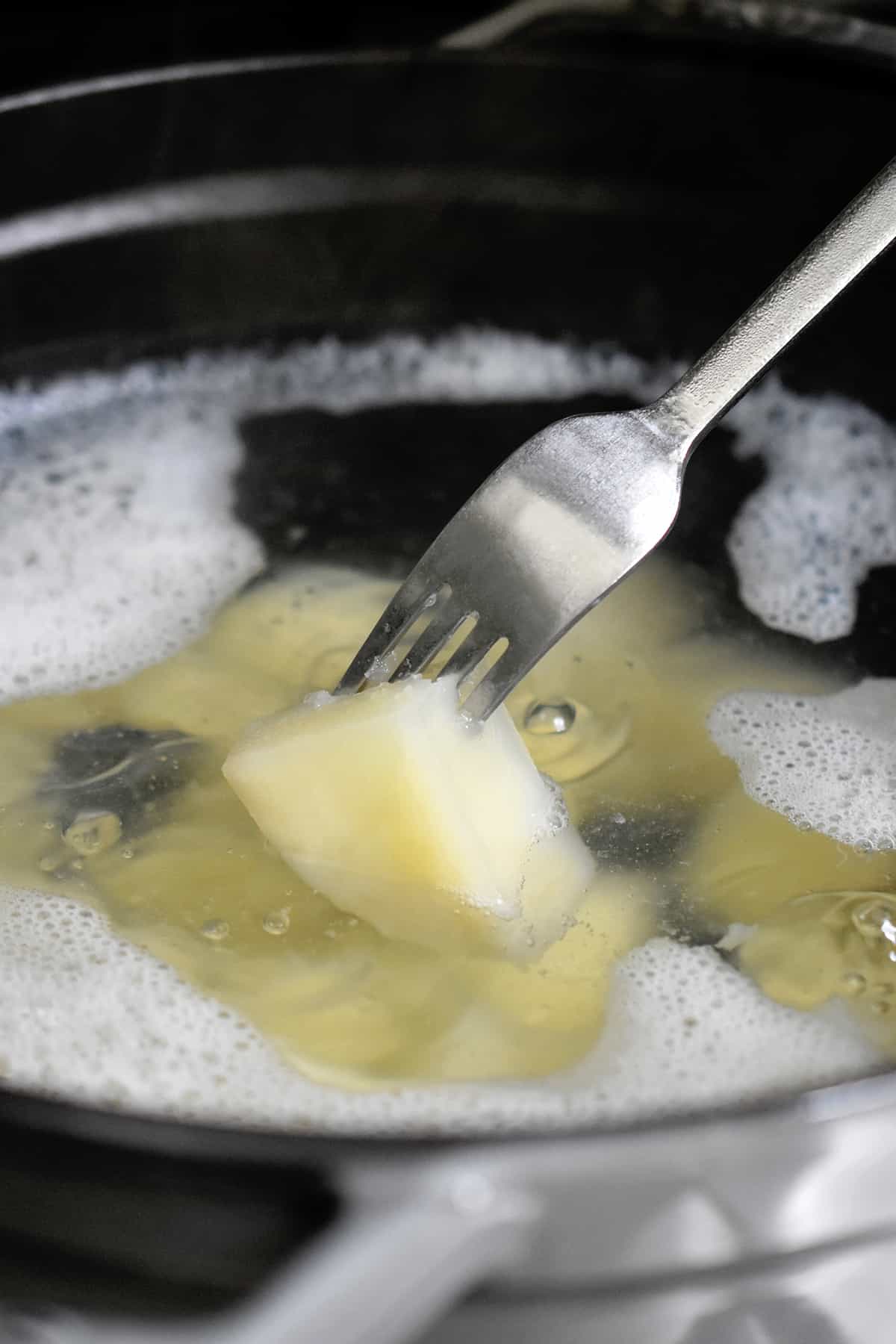 A fork is piercing a potato cube in a pot of boiling water.