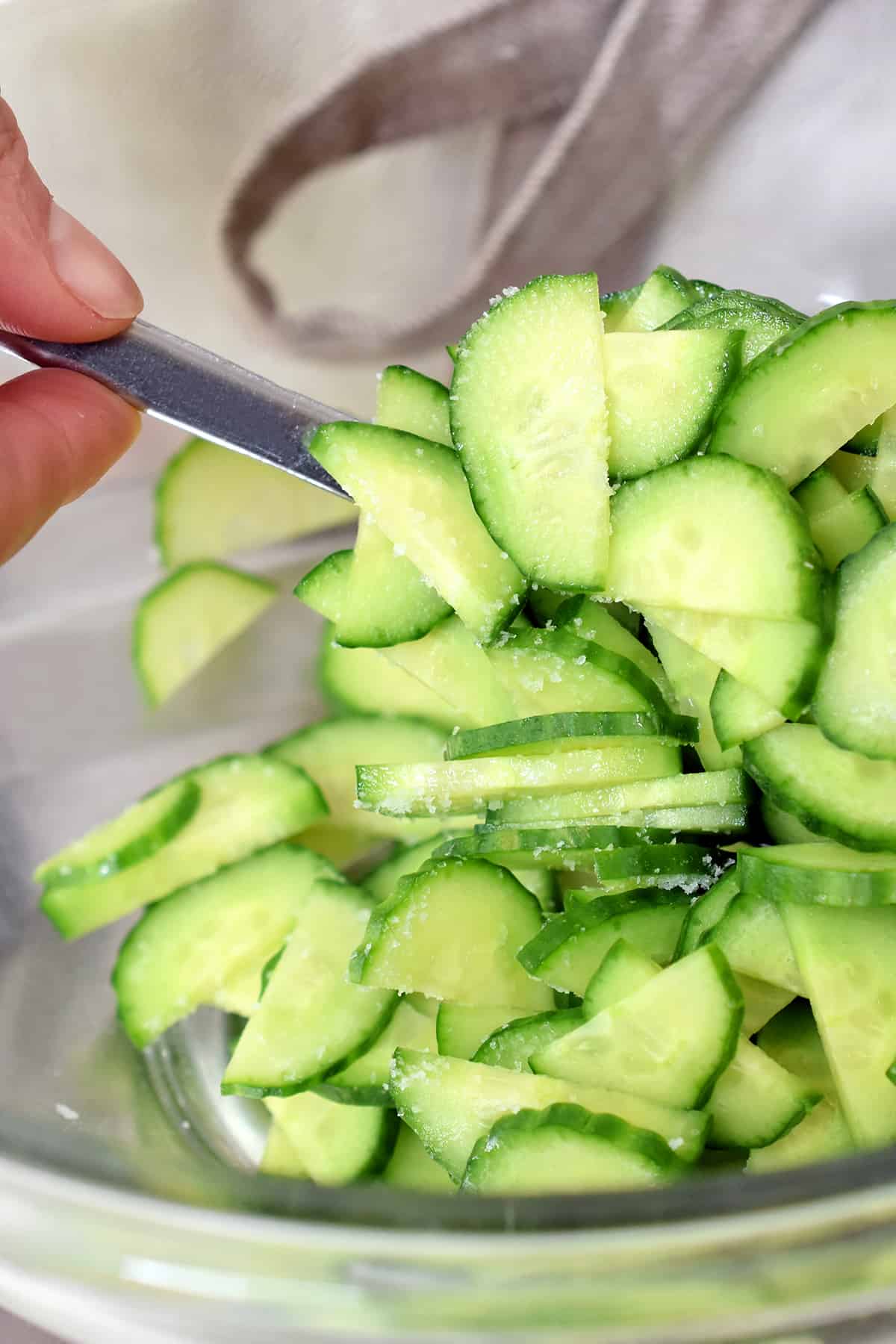 A closeup of sliced Persian cucumbers with salt sprinkled on top in a glass bowl.