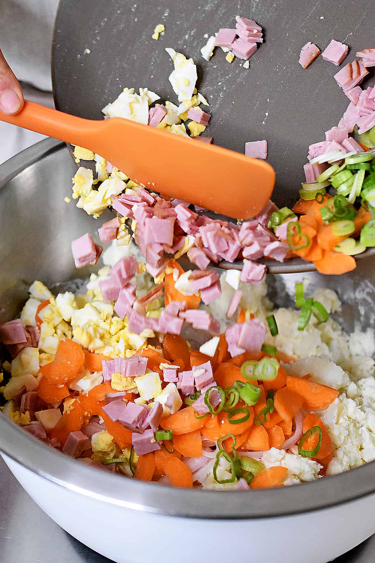Adding diced ham, sliced carrots, hard boiled eggs, and sliced green onions to a bowl filled with mashed potatoes to make Japanese potato salad.