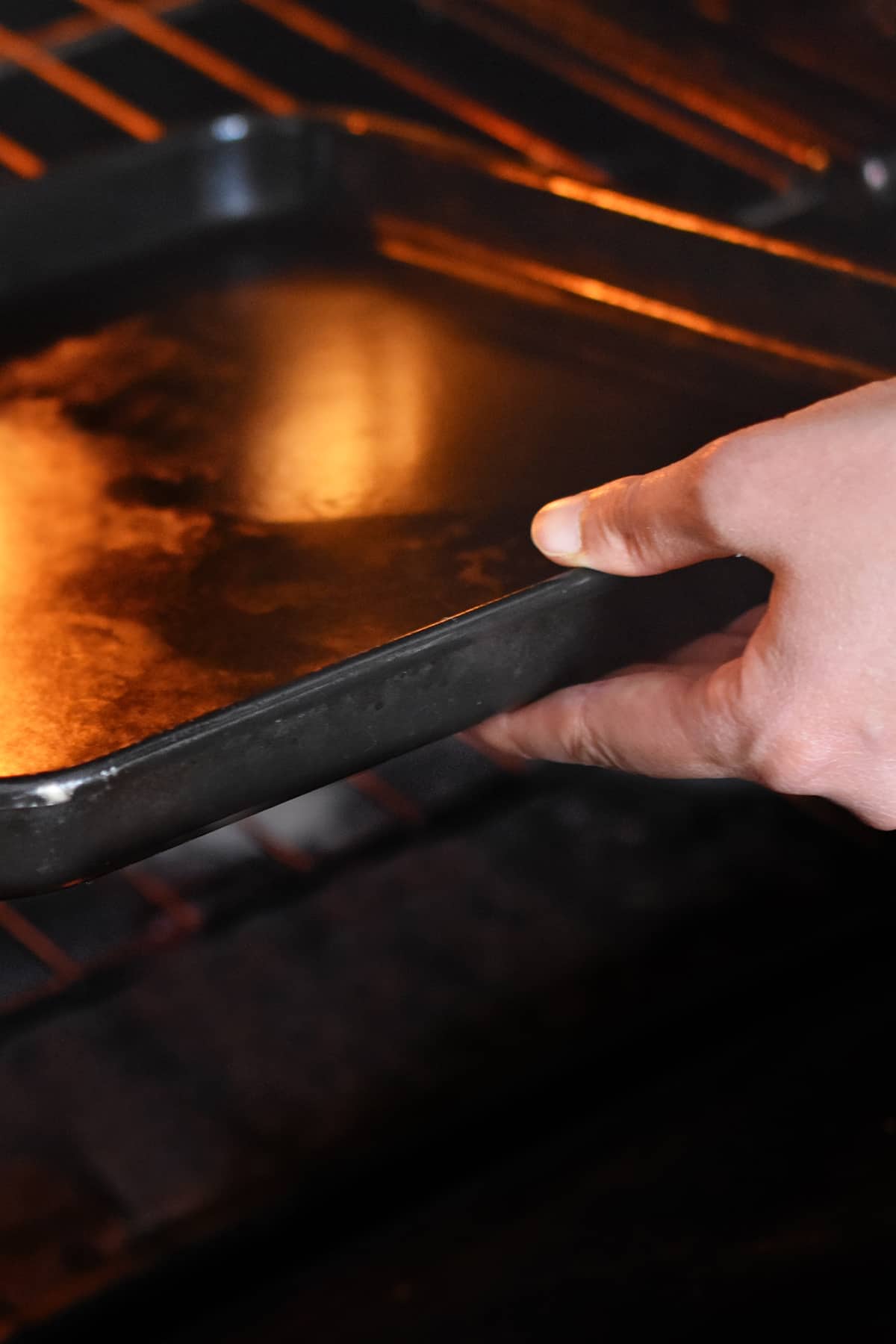 A hand is placing a black rimmed baking sheet into an open oven.