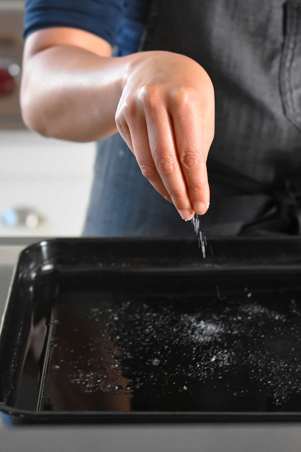 A hand is sprinkling salt into a black rimmed baking sheet with melted fat inside.