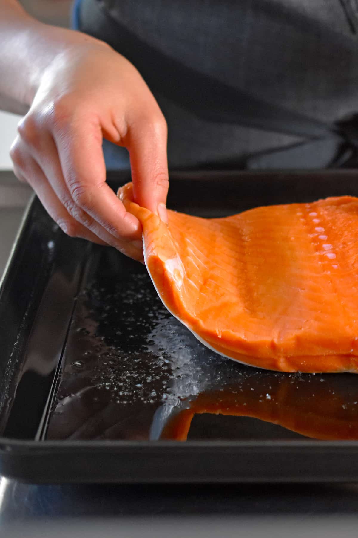 A hand is placing a raw salmon fillet, skin-side down, on a black rimmed baking sheet with melted ghee and salt.