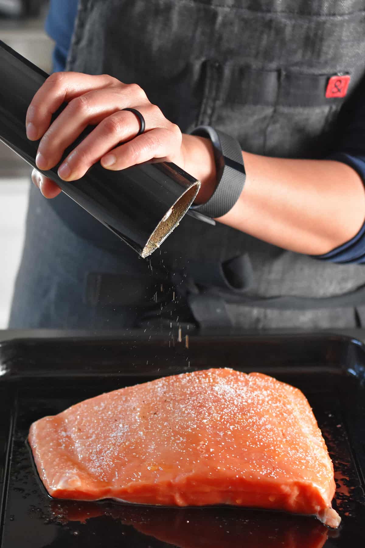 Adding black pepper from a pepper mill to a raw piece of salmon on a black rimmed baking sheet.
