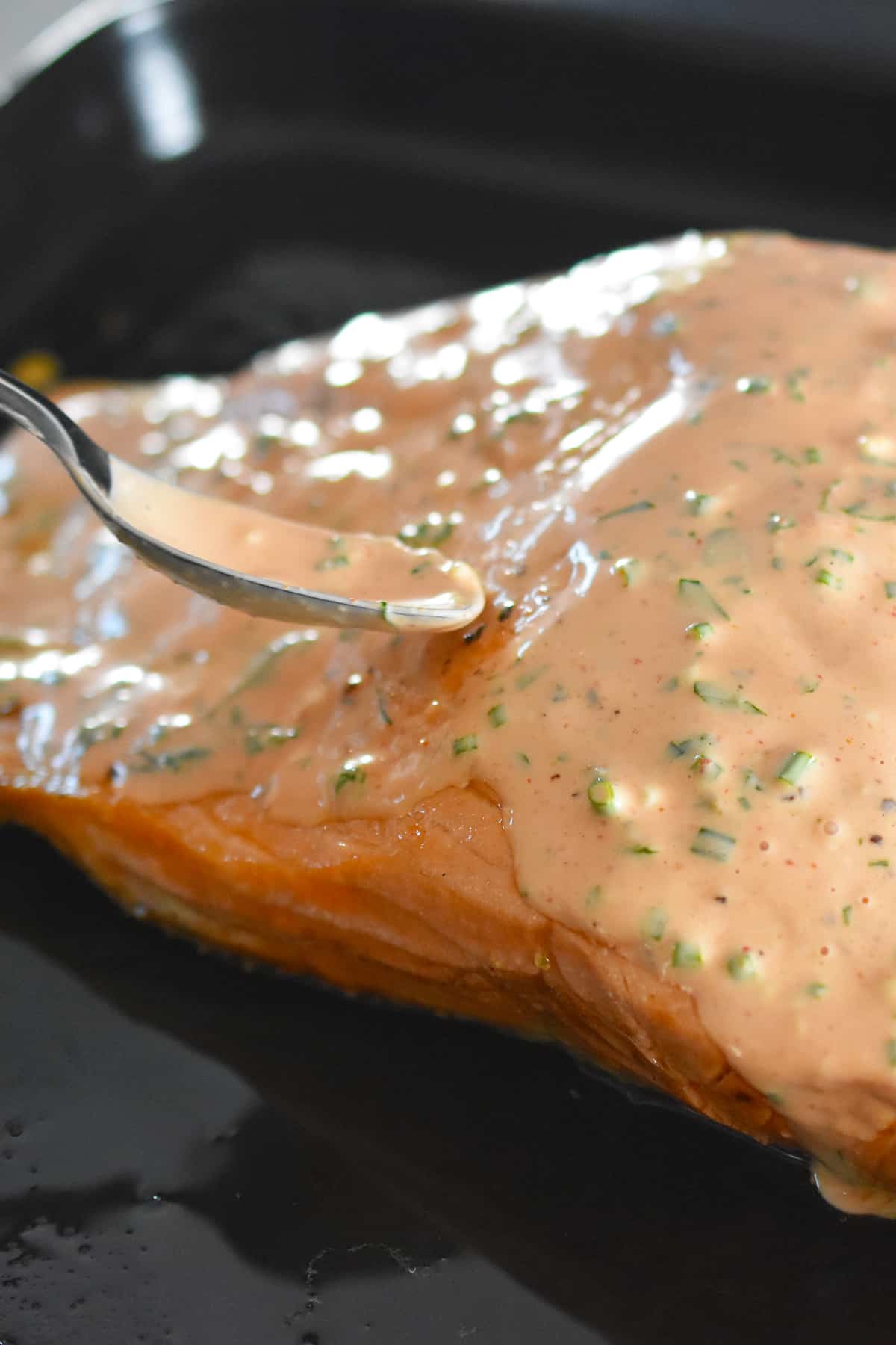 Spooning sriracha ranch dressing on a cooked salmon fillet in a black rimmed baking sheet.