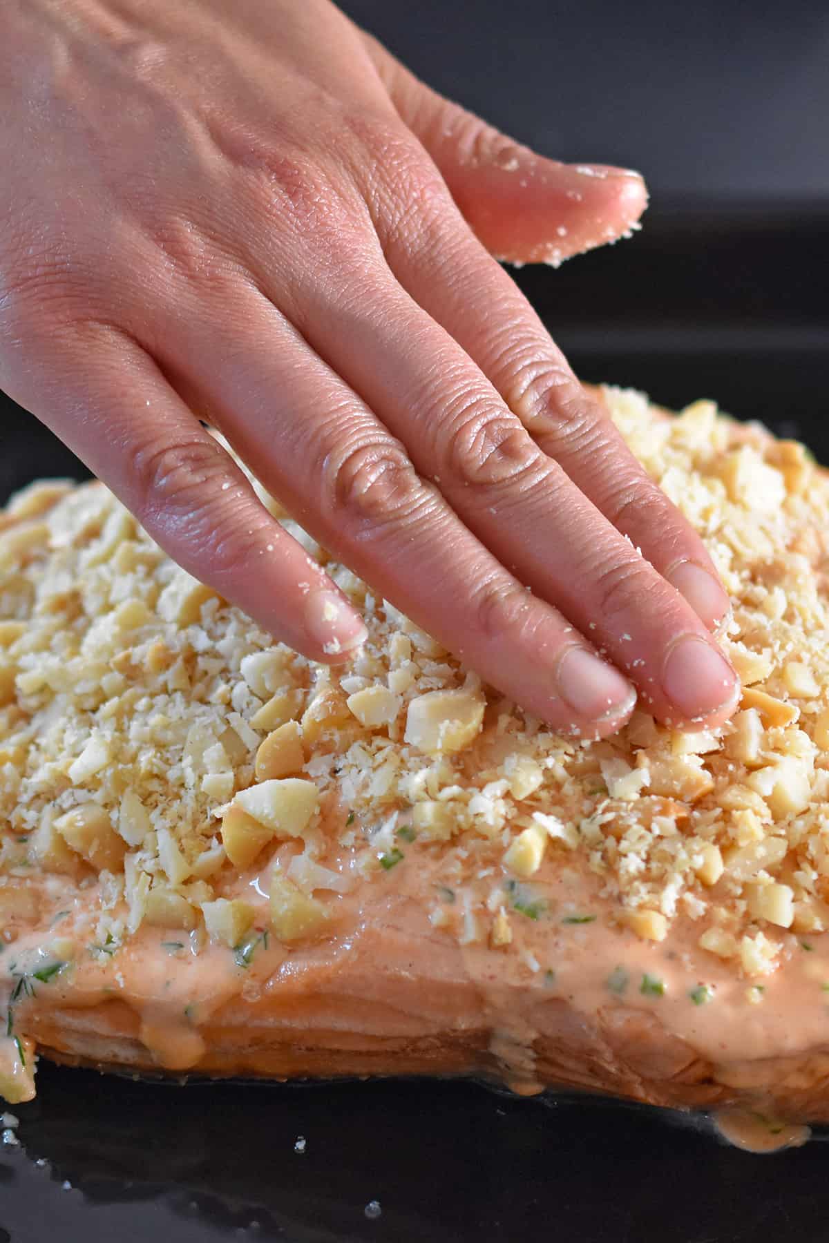A hand is applying chopped macadamia nuts to the top of a cooked salmon fillet that is coated with sriracha ranch dressing.