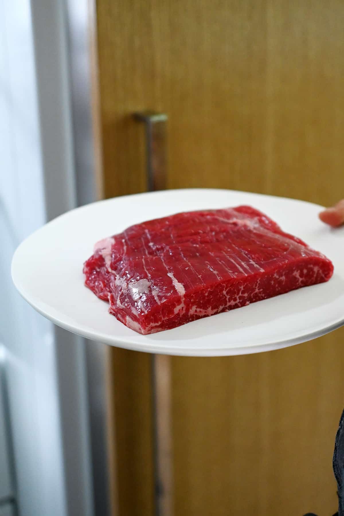 A hand is holding a white plate with a raw piece of flank steak that is headed for the freezer.