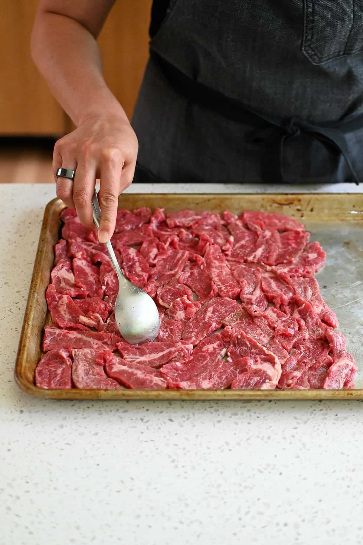 A spoon is arranging sliced raw flank steak in a single layer on half of a sheet pan.