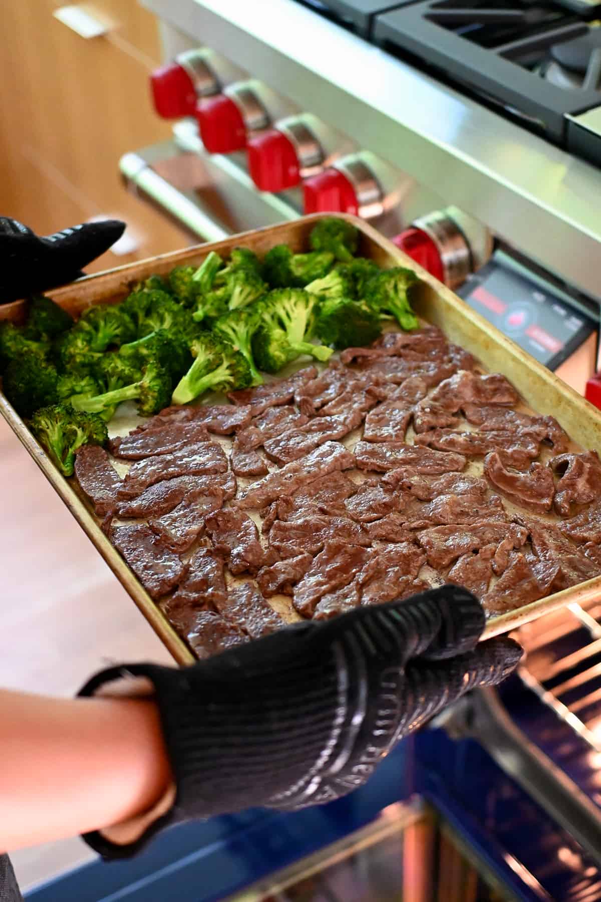 Someone lifting a sheet pan filled with sliced beef and broccoli out of the oven so sauce can be added to it.
