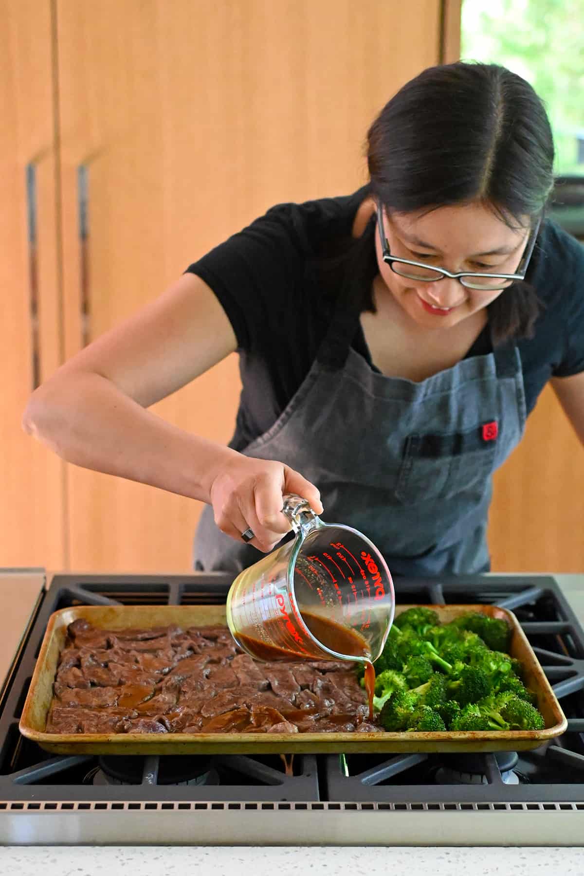 An Asian woman is pouring a brown sauce onto a sheet pan filled with sliced beef and broccoli.
