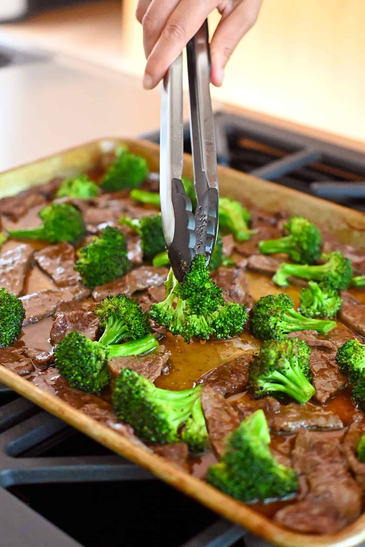A pair of tongs is rearranging the beef and broccoli on the sheet pan before putting it under the broiler.