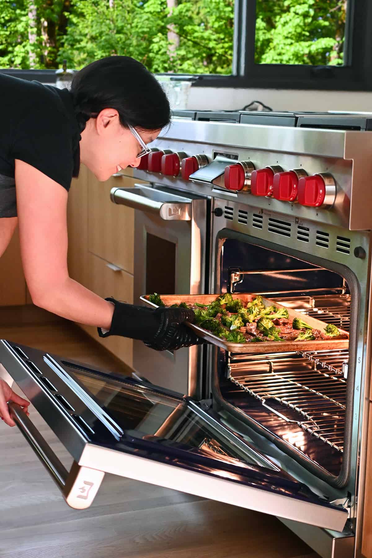 An Asian woman is putting a sheet pan beef and broccoli under the broiler in an open oven.