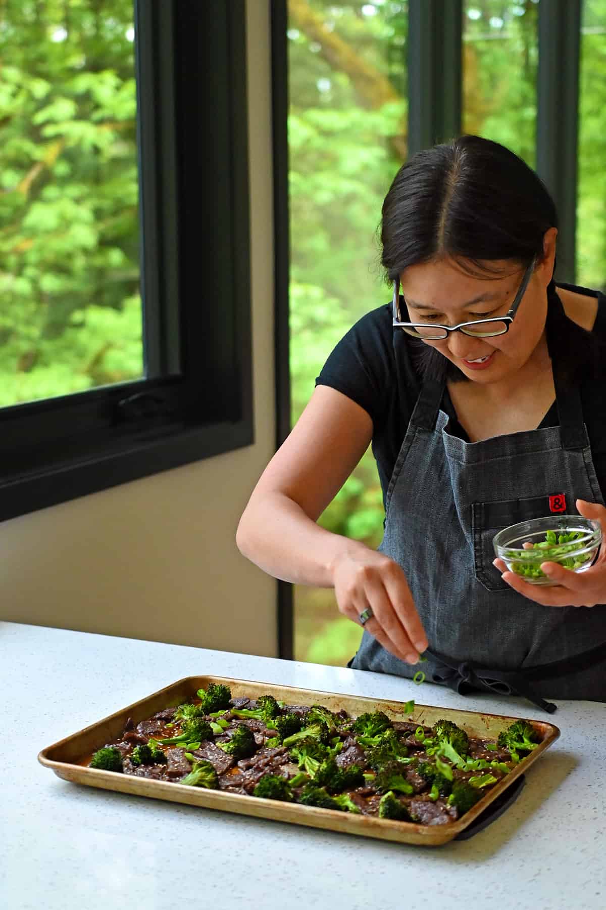 An Asian woman is sprinkling sliced green onions onto a rimmed baking sheet filled with Chinese beef and broccoli.