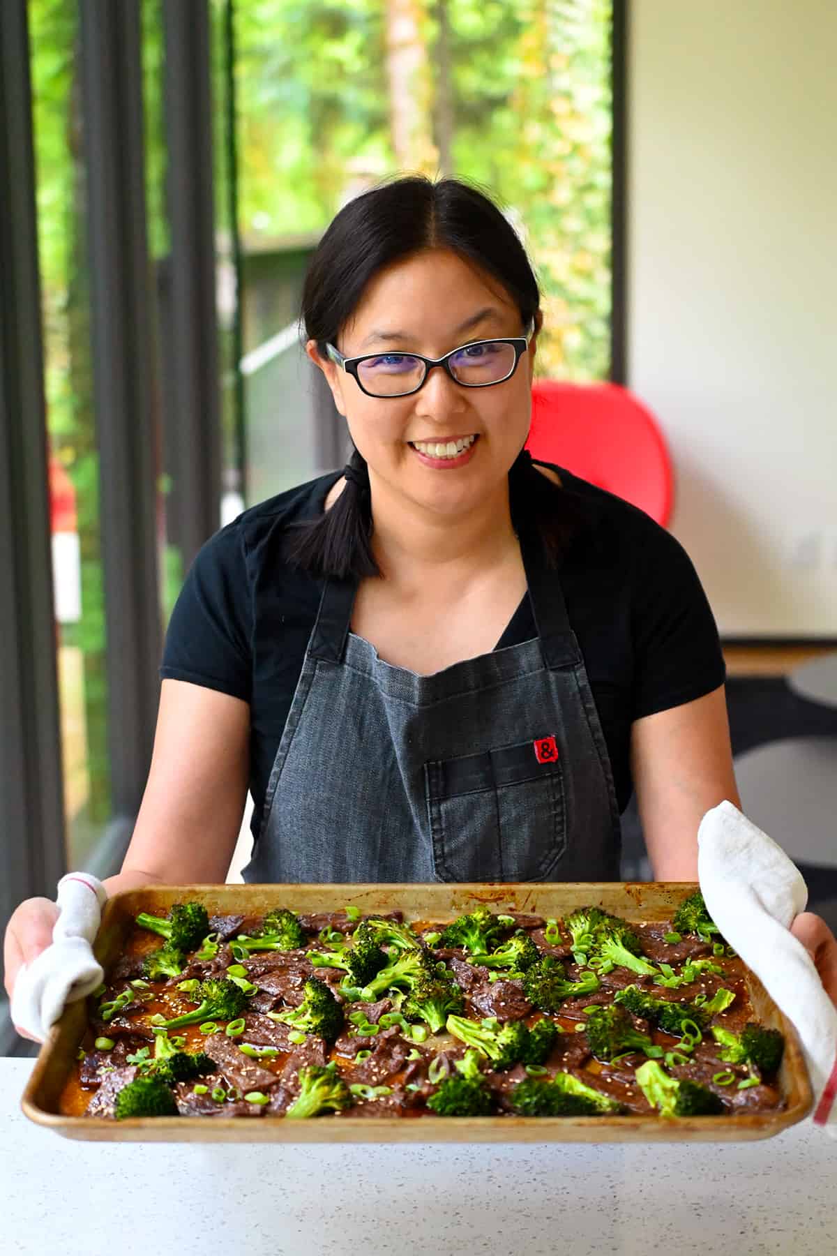 A smiling Asian woman is holding a sheet pan filled with Chinese beef and broccoli right out of the oven.