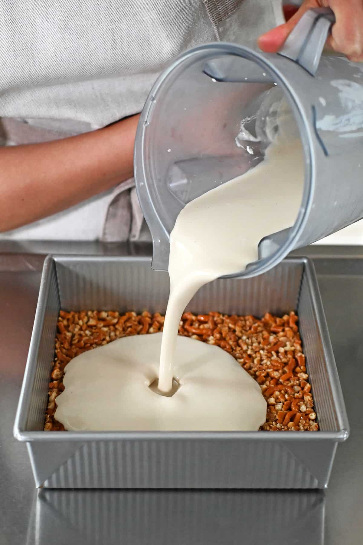 Pouring a creamy filling from a blender onto a grain-free pretzel crust in a square baking pan.