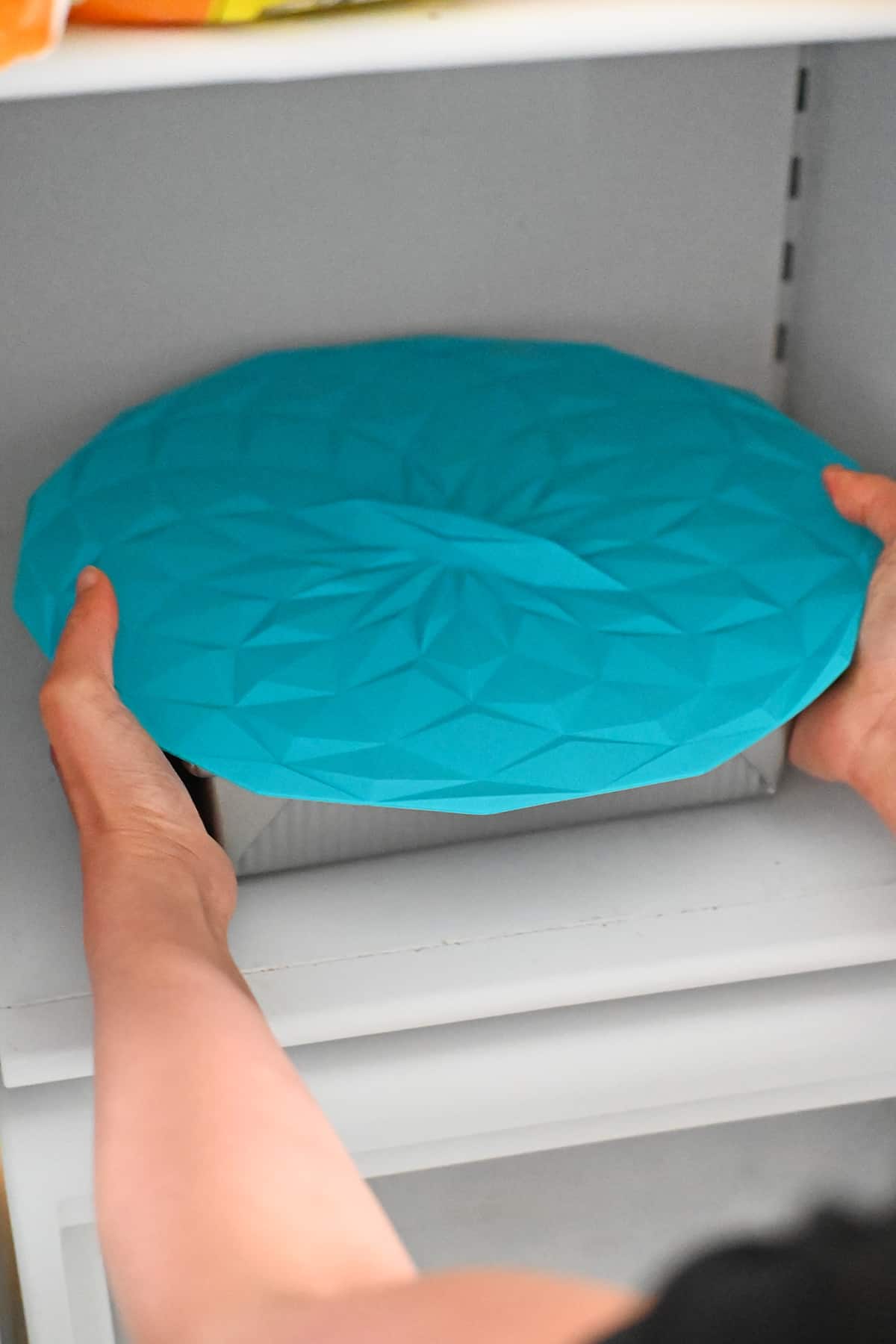 A square baking pan filled with strawberry pretzel salad is covered with a blue silicone lid as it is being placed in a freezer.