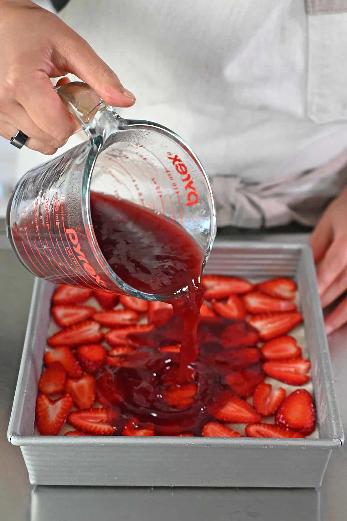 Pouring homemade strawberry gelatin from a liquid measuring cup on top of a strawberry pretzel salad in a square baking pan.