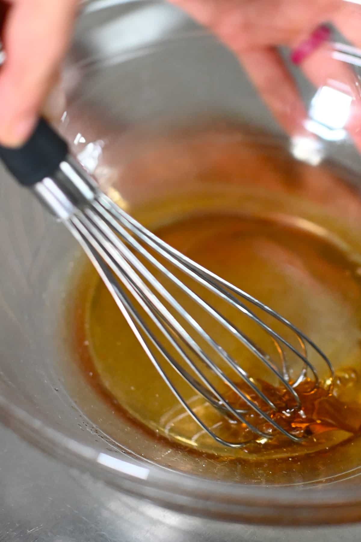 Closeup of a whisk mixing melted coconut oil and maple syrup in a clear mixing bowl.