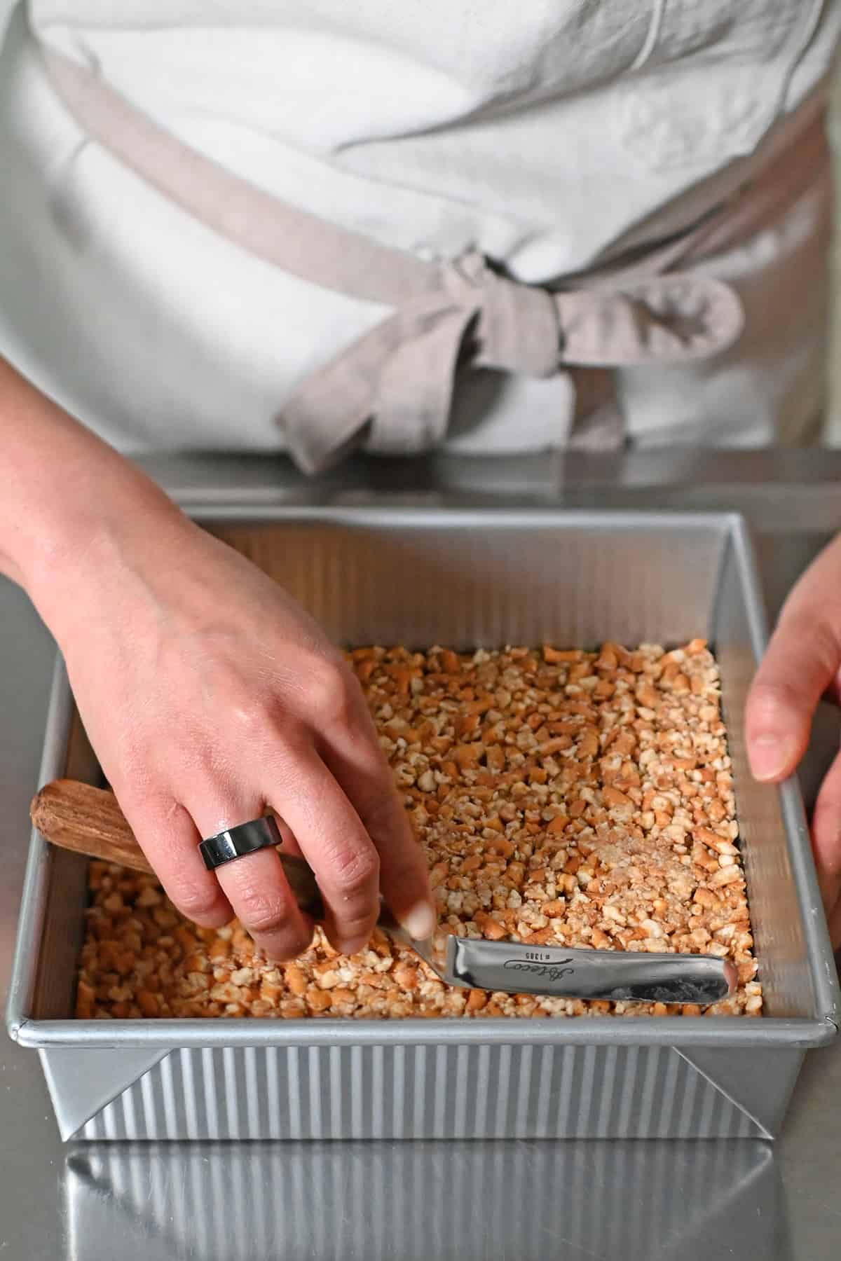 A hand is using a small offset spatula to press the pretzel crust into an even layer on the bottom of a square baking pan.