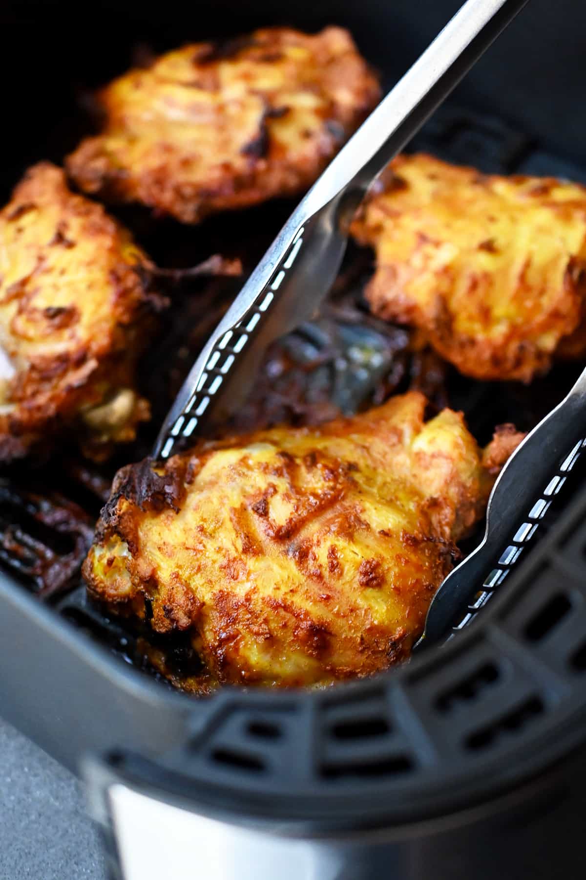 A pair of tongs is grabbing a golden brown tandoori chicken thigh from an open air fryer.