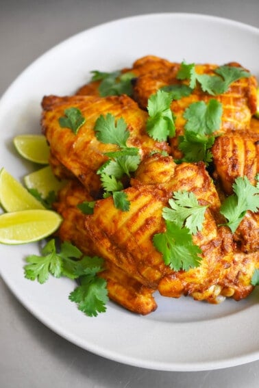 A plate with tandoori chicken thighs topped with cilantro leaves and some lime wedges.