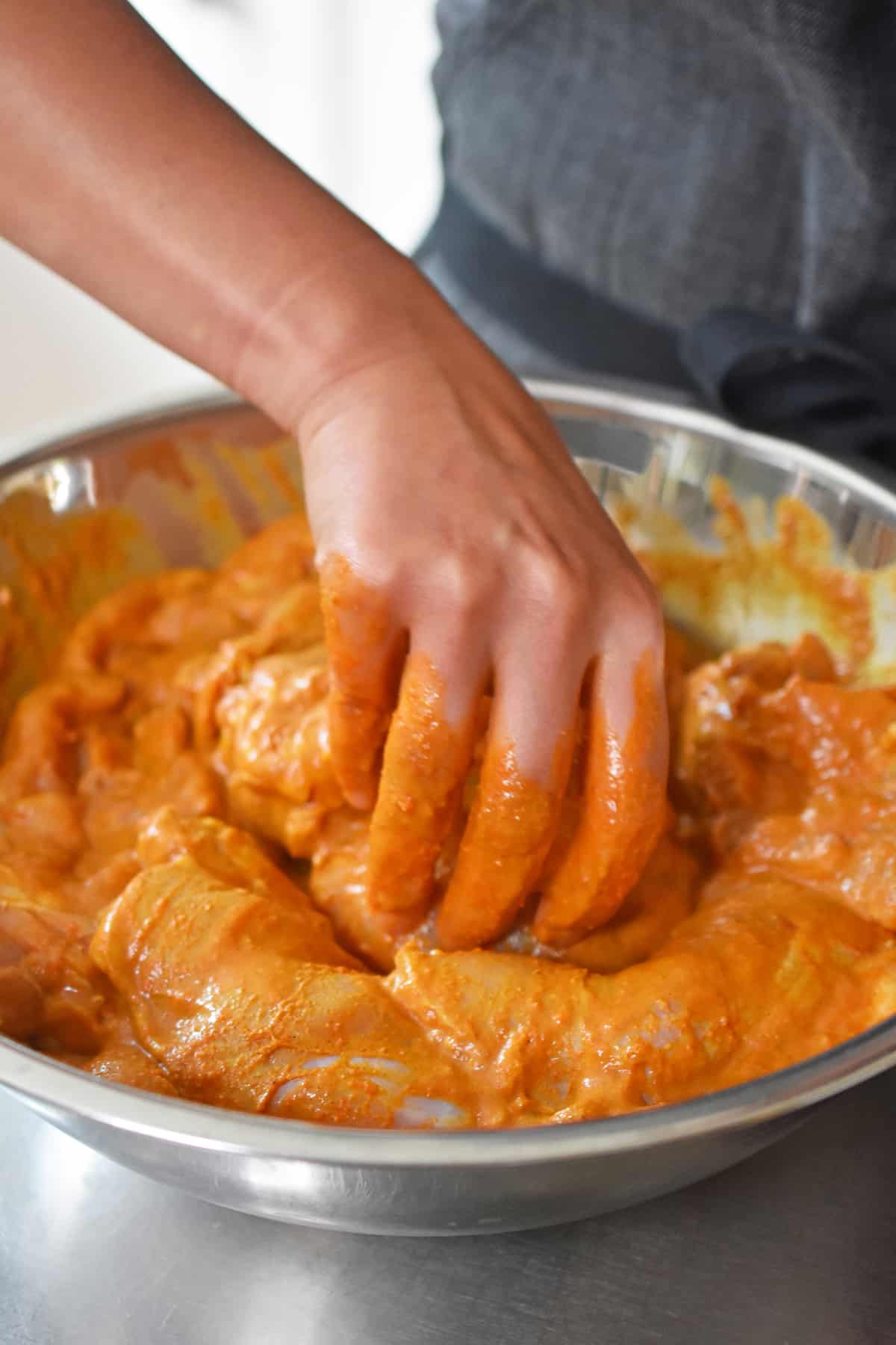 A hand is tossing raw chicken thighs in a bright orange colored tandoori marinade.