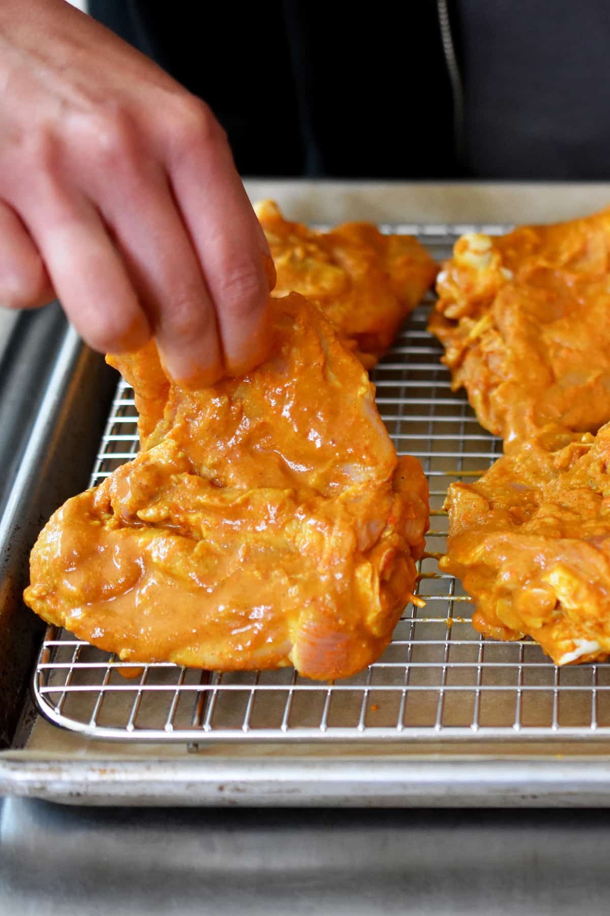 A hand is placing a raw piece of tandoori chicken thigh on a wire rack in a rimmed baking sheet.