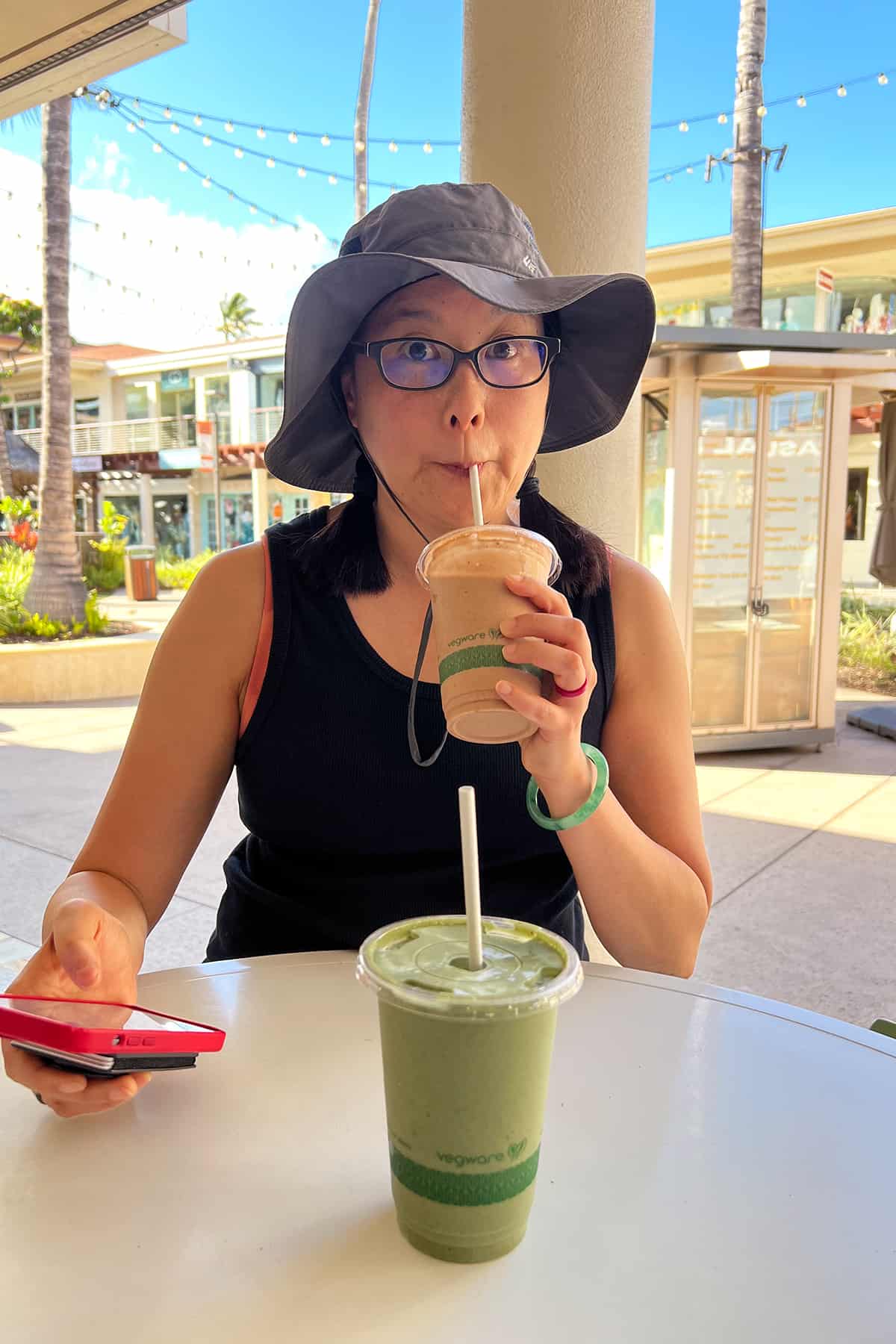 An Asian woman in a hat is drinking a brown smoothie while holding a red cell phone.