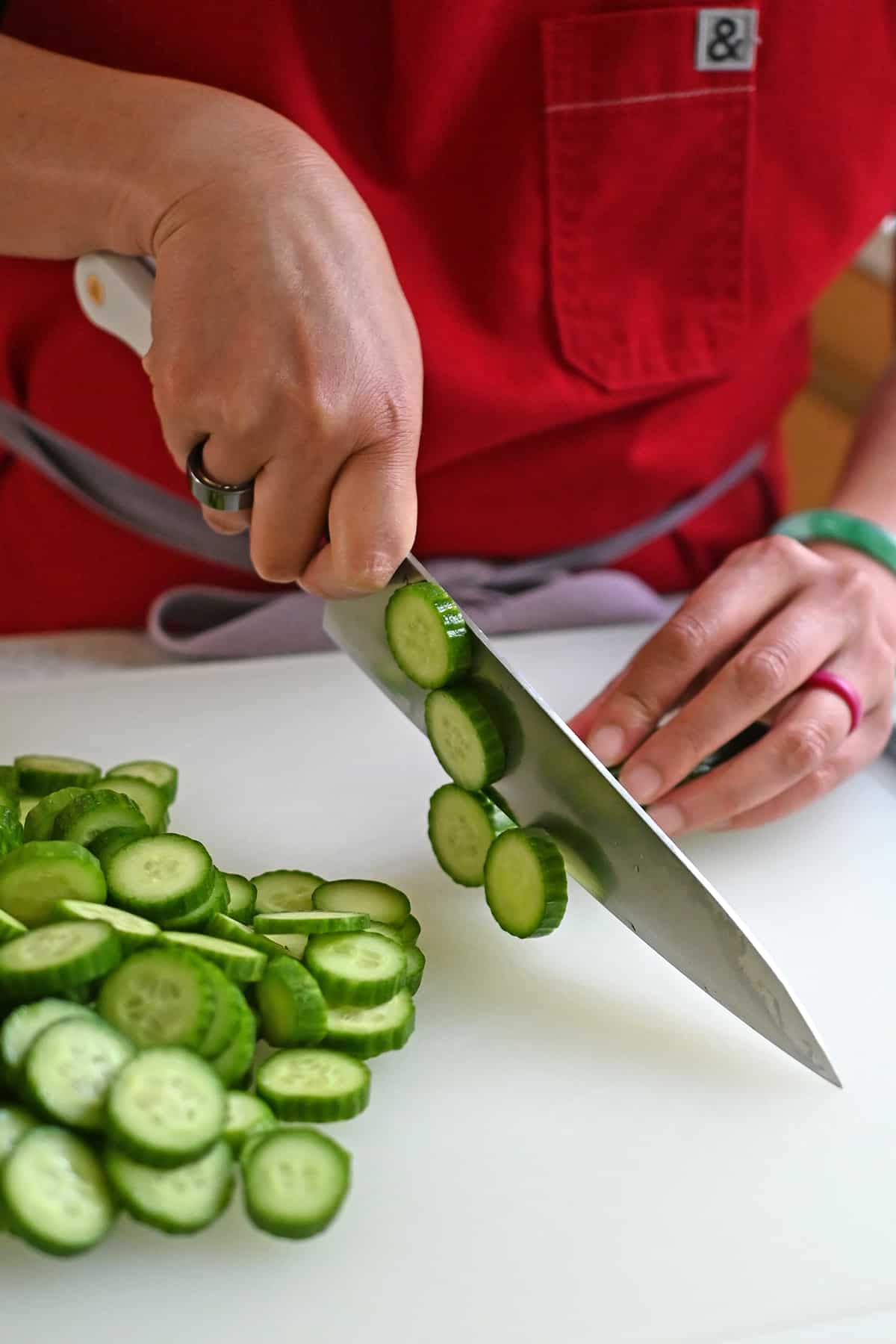 A person in a red apron is cutting Persian cucumbers into thin slices with a chef knife.