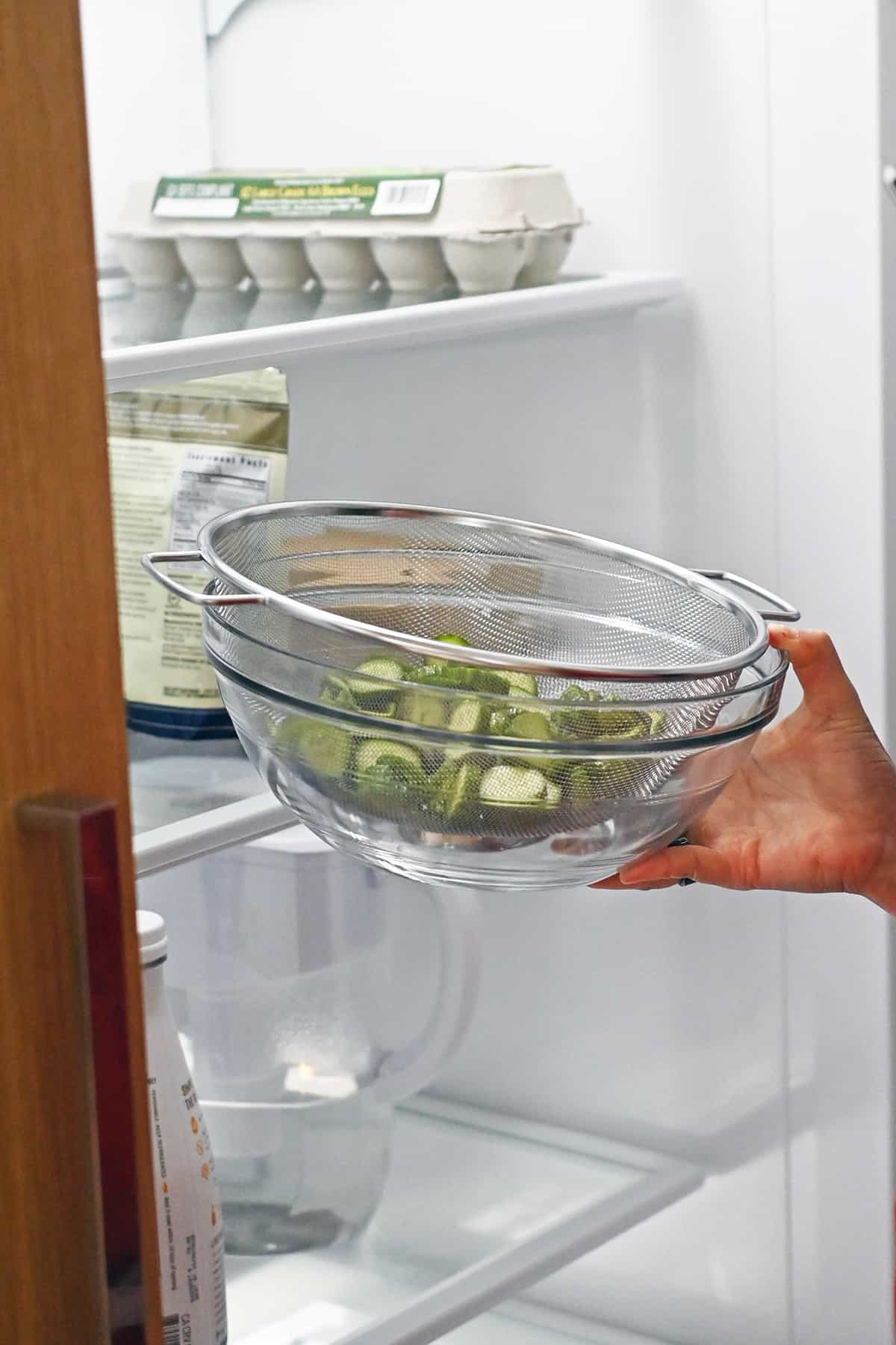 Placing salted cucumber slices in a colander and glass bowl into the refrigerator.