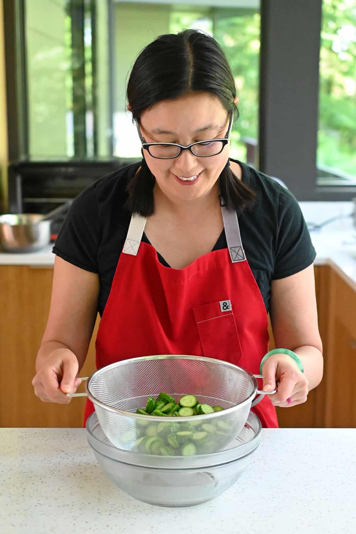 Placing a colander filled with salted sliced cucumbers into a large glass mixing bowl.