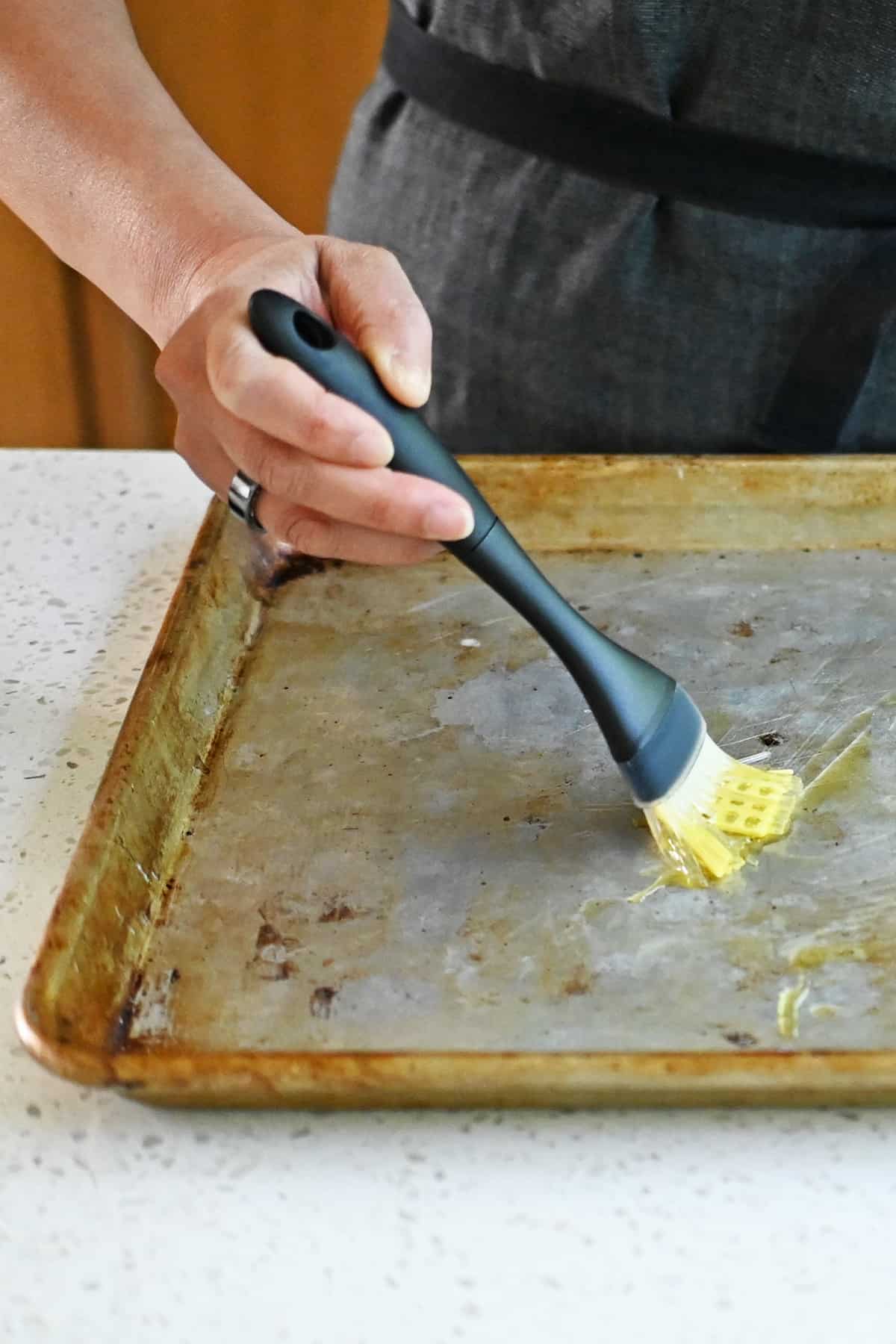 A silicone brush is spreading extra virgin olive oil evenly on a rimmed baking sheet.