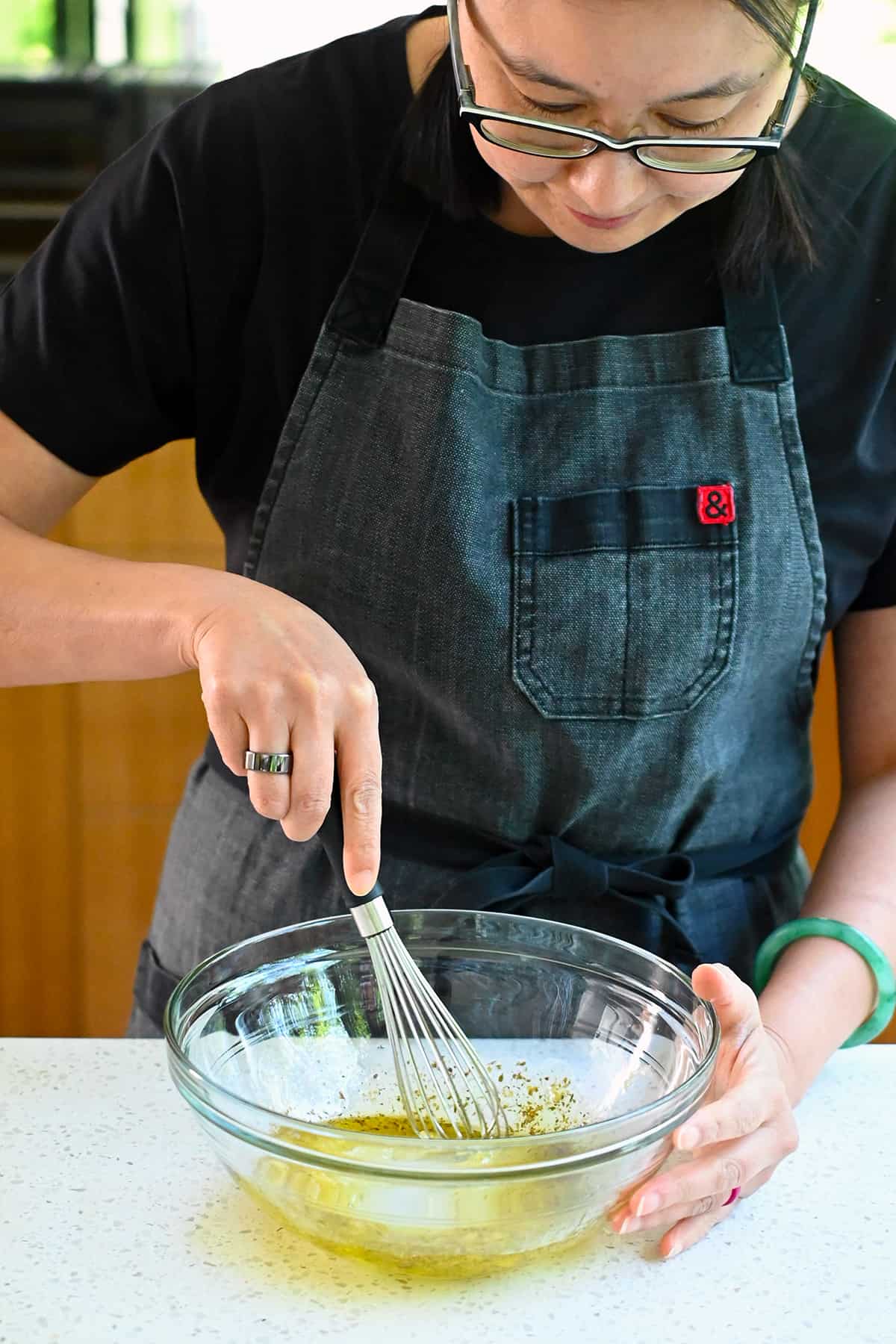 An Asian woman is whisking a Greek marinade in a clear glass mixing bowl.
