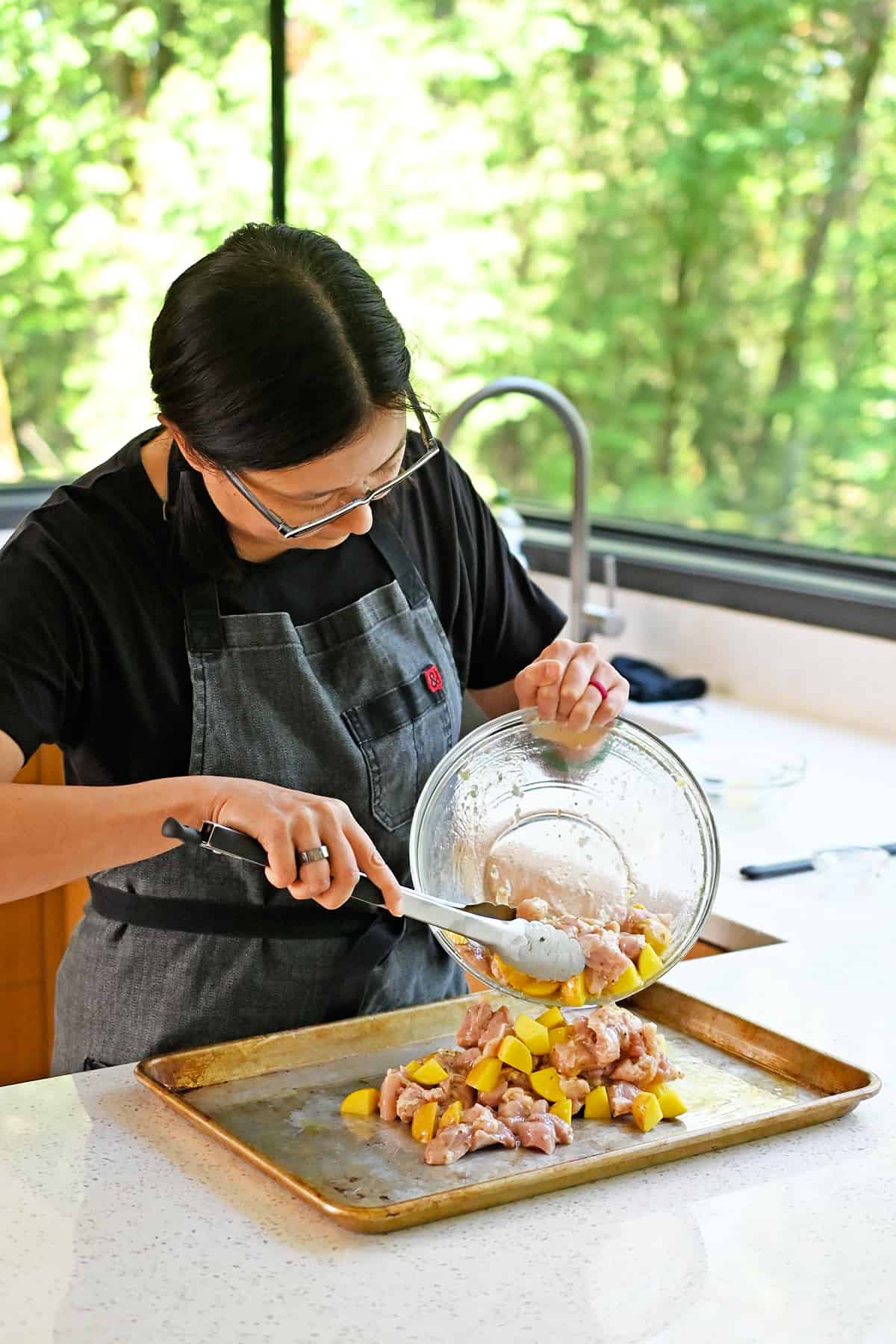 An Asian woman is transferring the marinated raw chicken and potatoes onto a greased rimmed baking sheet.