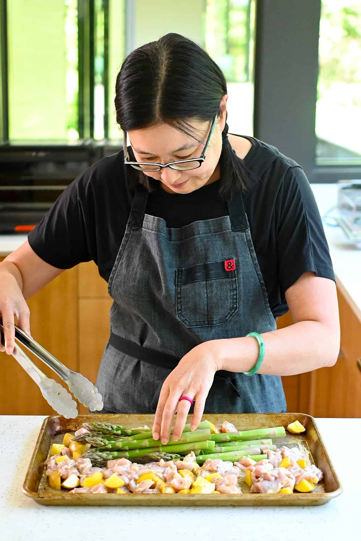 An Asian woman is placing some asparagus spears on a sheet pan with chicken and potatoes marinated with a Greek dressing.