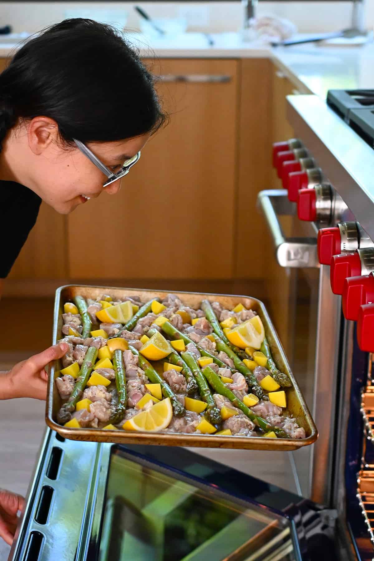 An Asian woman is placing a Greek sheet pan chicken dinner into an open oven.